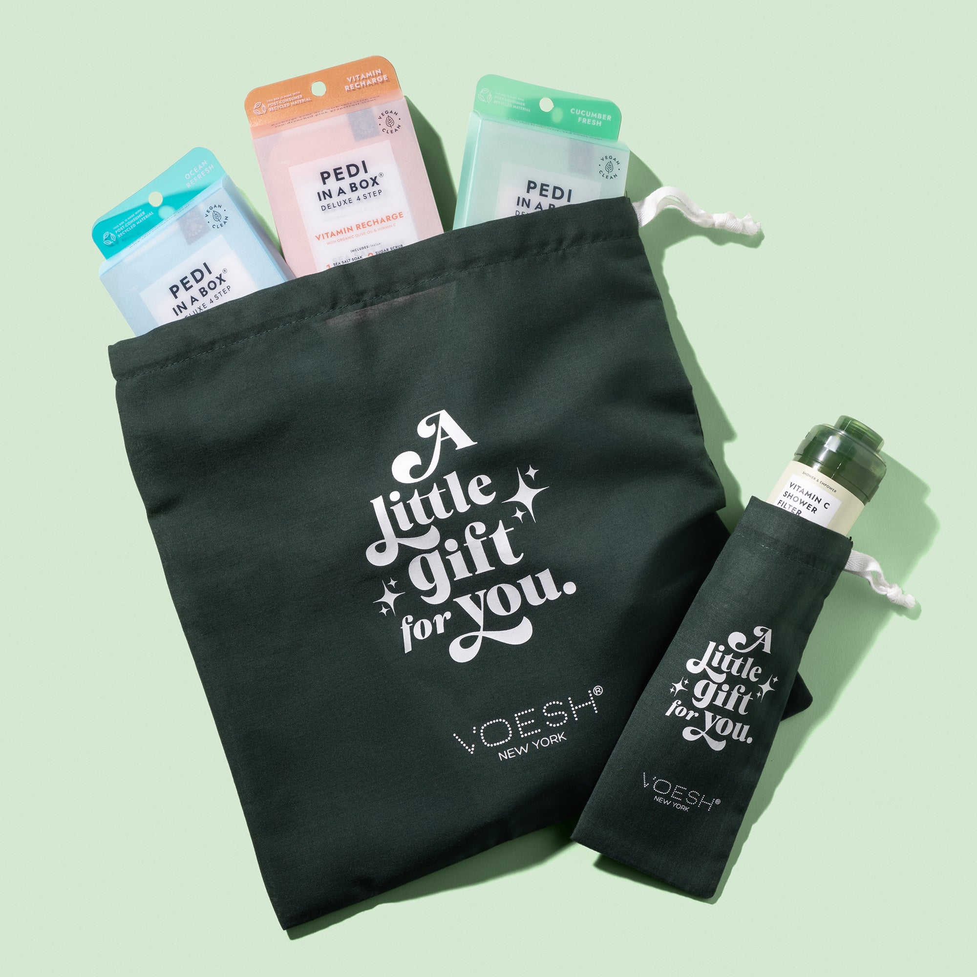 Two VOESH Drawstring Gift Bags, one small and one large, stuffed with VOESH products on green background.